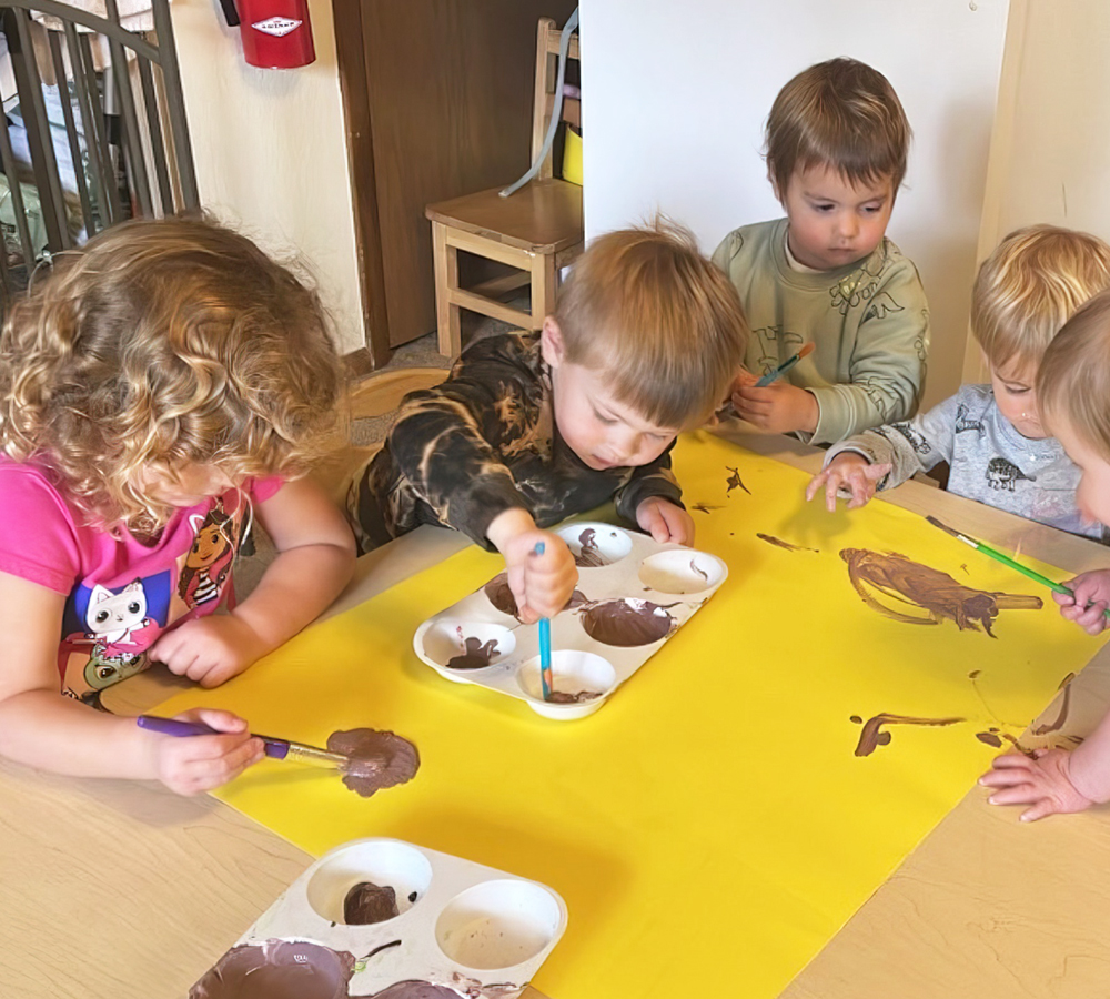 A group of five children focused on painting at a table, each using brushes and colorful materials, engaged in creative play