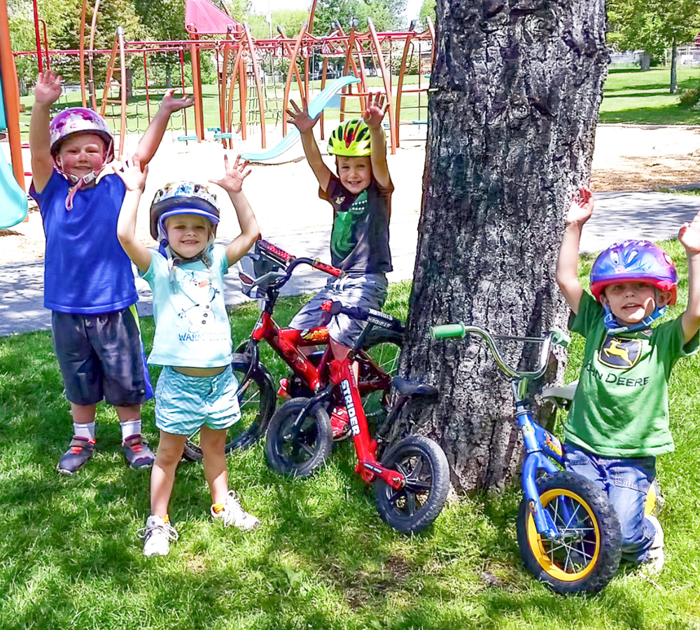 A group of four children in helmets, excitedly raising their arms near their bicycles in a sunny park, expressing joy.
