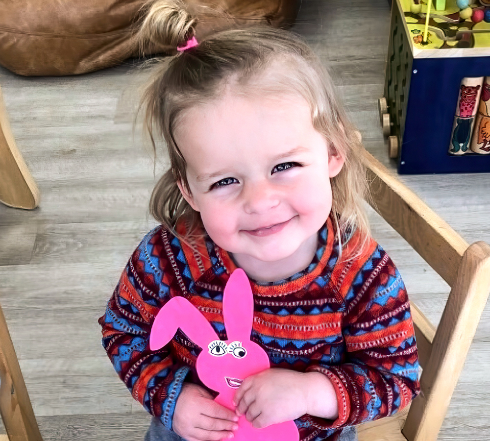 A young girl holding a pink paper bunny, smiling brightly, with a playful and cheerful expression.