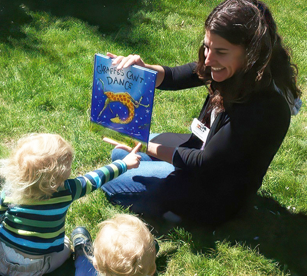 A teacher reading "Giraffes Can't Dance" to two children sitting on grass, engaging them with storytelling.