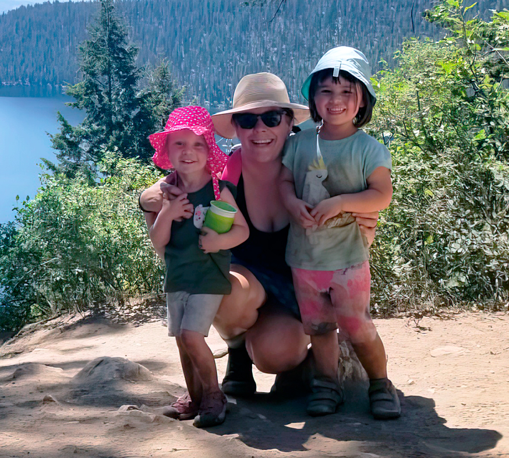 Teacher kneels between two smiling children, with trees and mountains in the scenic background.