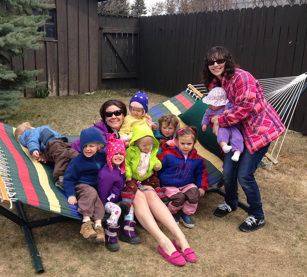 A group of smiling kids and teachers relax together on a hammock, enjoying a sunny outdoor day.