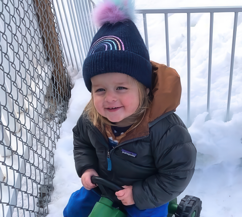 A cheerful child sits on a toy ride-on in the snow, wearing a cozy hat and a big smile.