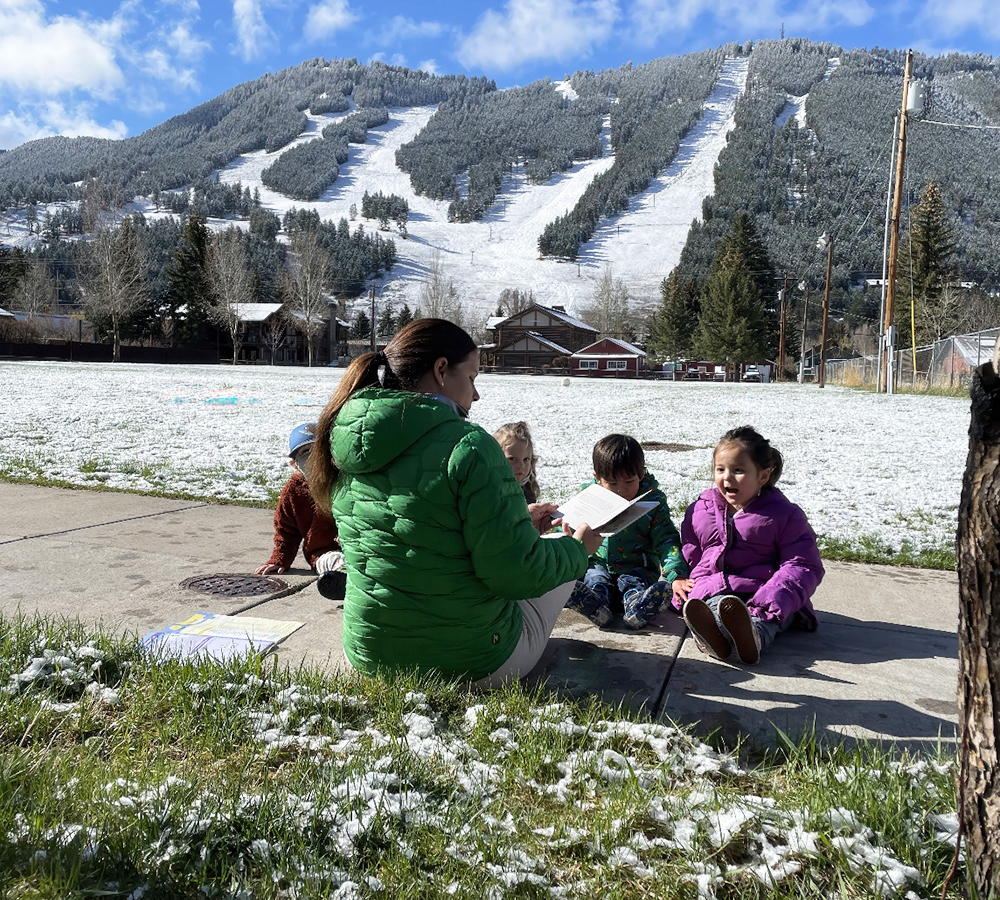 A teacher engages with children sitting outside in a snowy area, sharing stories and educational moments together.