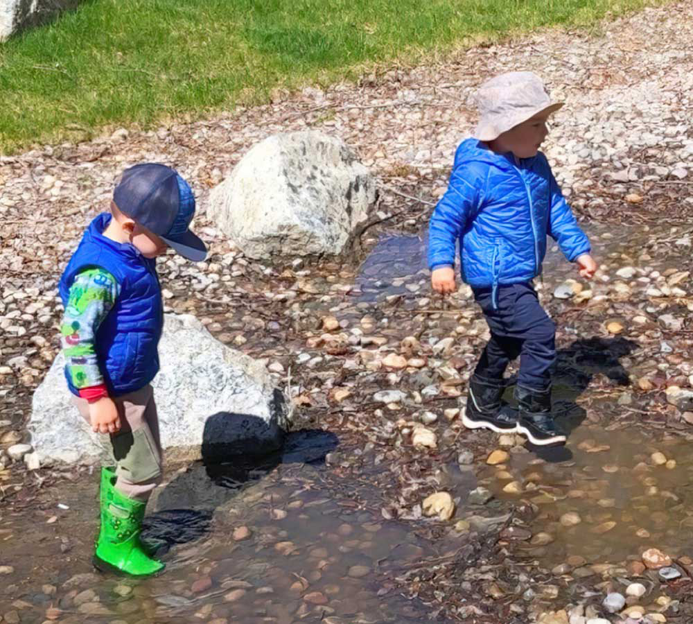 Two children walking through shallow water on a rocky shore, wearing jackets and exploring nature