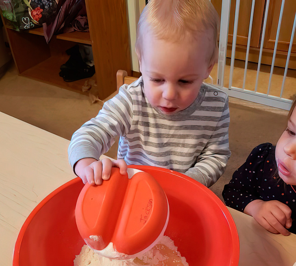 2 kids are happily mixing ingredients in a bowl, showing curiosity and excitement, while another child watches nearby.