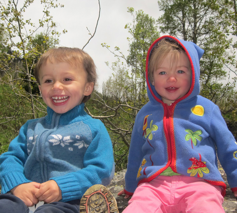 Two kids are sitting together outdoors, both smiling widely, dressed warmly, and enjoying their time in nature.