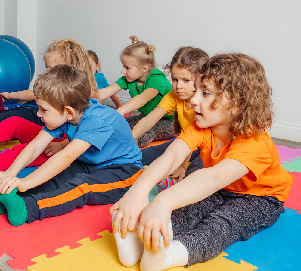 A group of children is participating in a stretching activity, sitting in a line on colorful mats, focused and engaged.