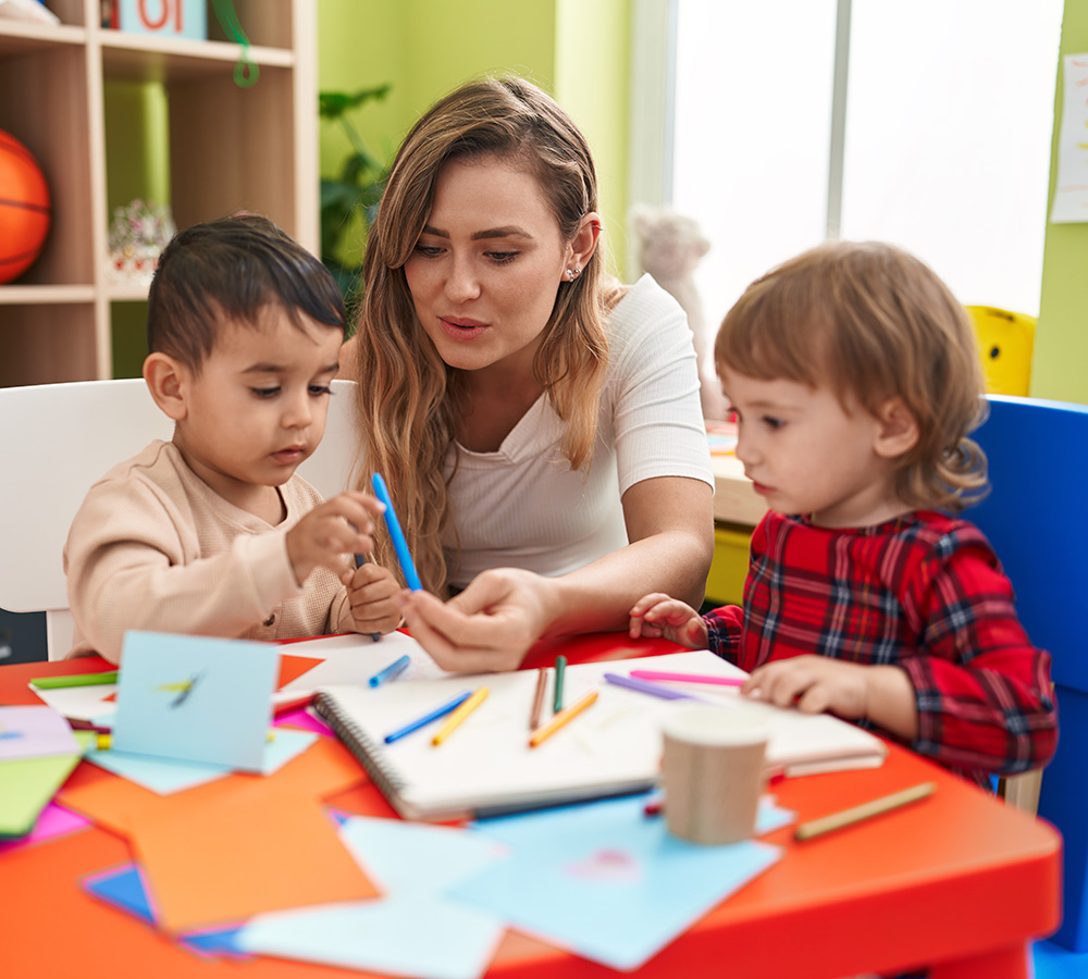 A teacher is helping two children with an arts and crafts activity, guiding them as they use colorful paper and markers.