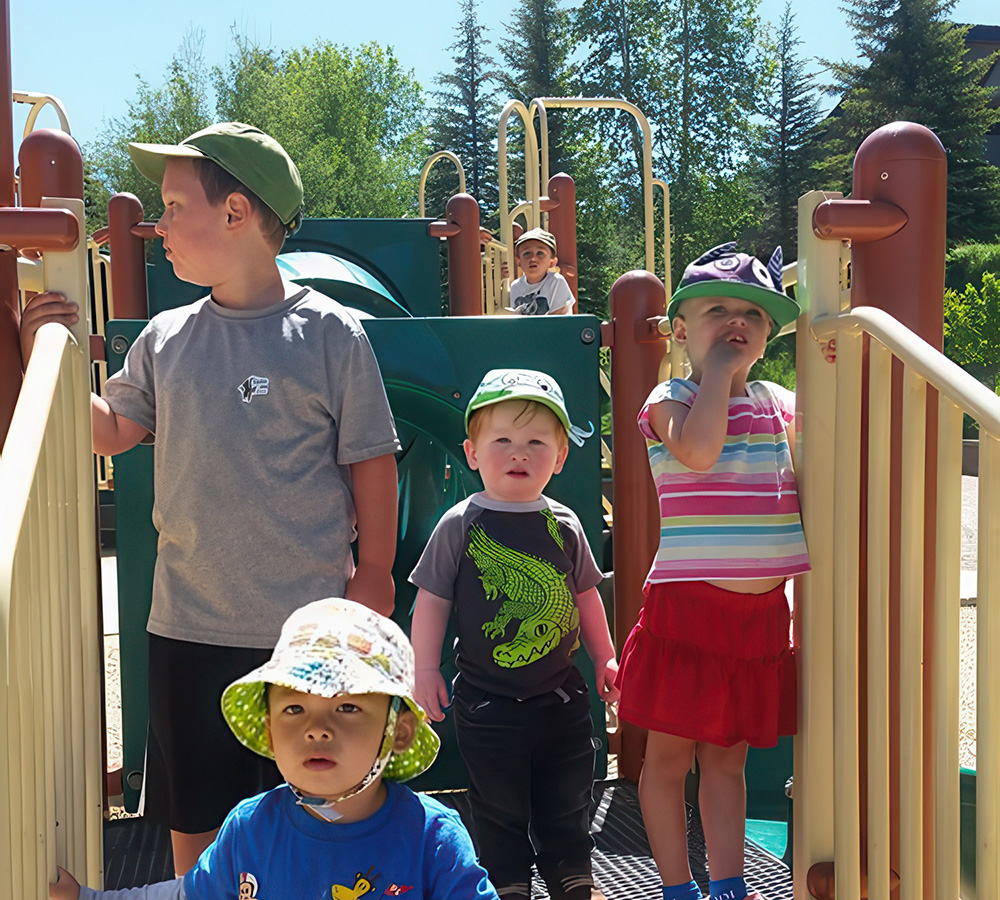 A group of children play on a playground, wearing hats and looking excited in the bright sunshine.