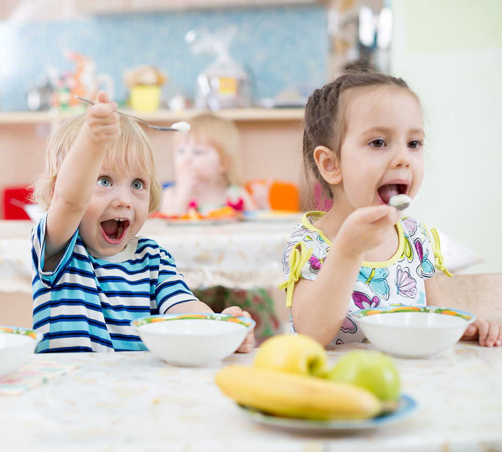 Two young children are happily eating from their bowls, smiling excitedly as they enjoy their meal in a playful setting.