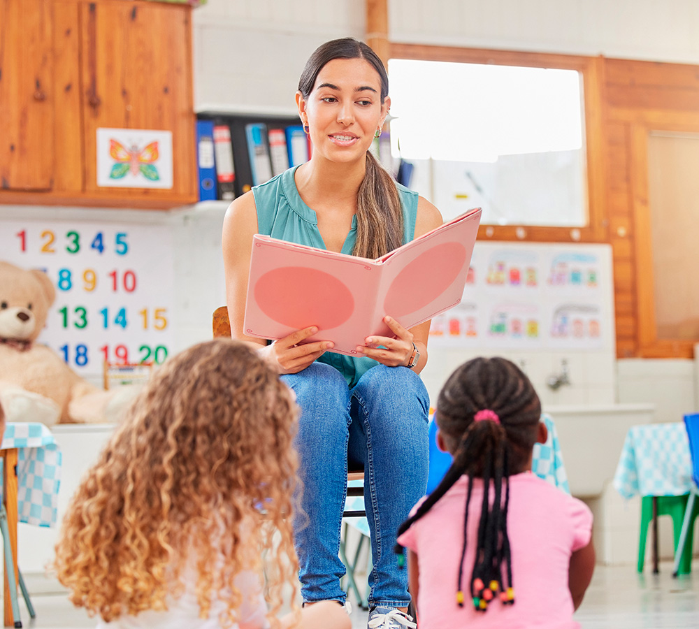 A teacher is reading a storybook in a group of children, engaging them with animated expressions.