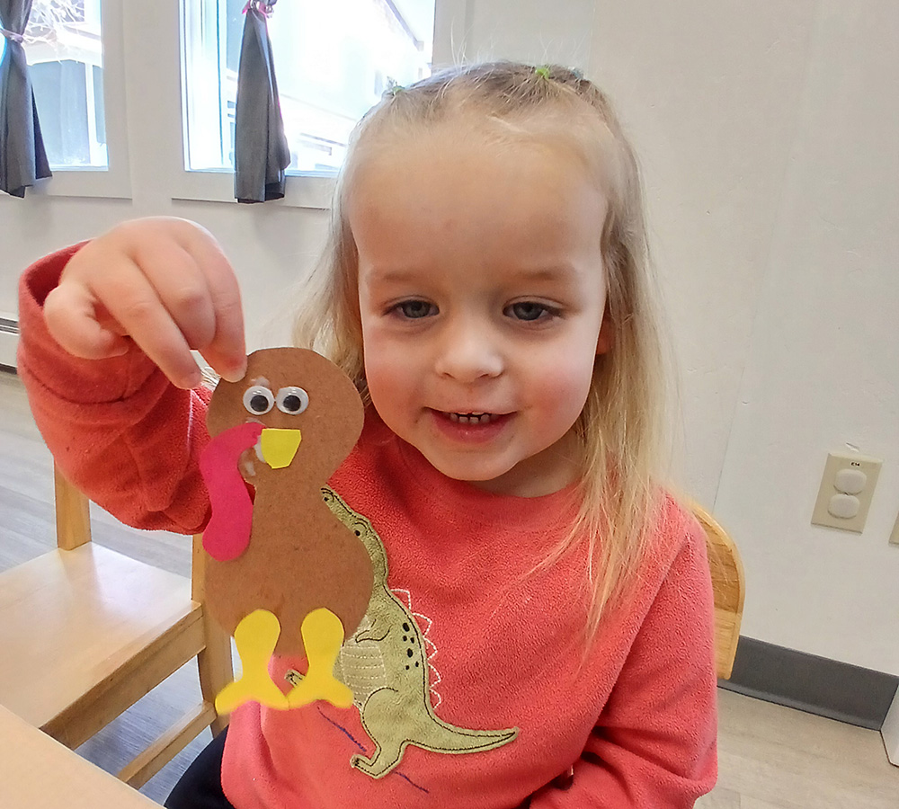 A girl is proudly holding up a handmade craft, featuring a colorful turkey, with a big smile on her face.