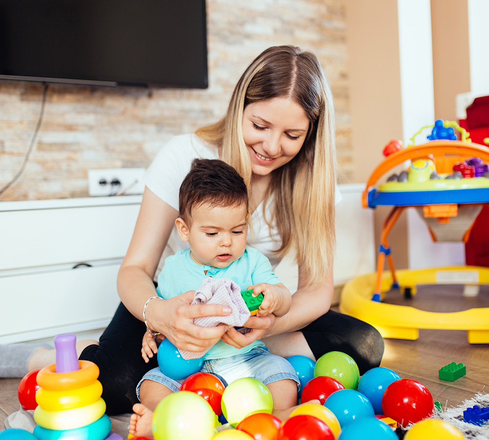 A teacher is playing with a baby, who is exploring colorful toys, both smiling and enjoying their time together.
