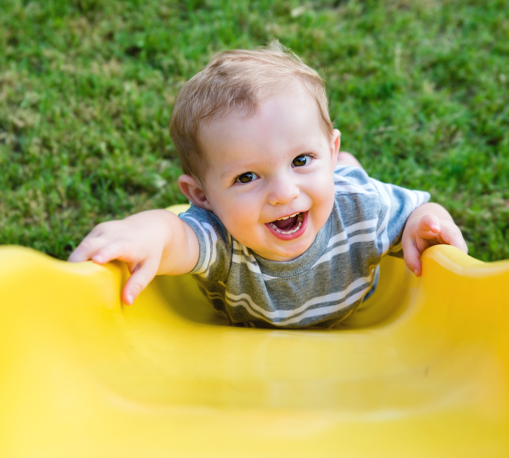 A smiling boy is climbing up a slide, looking excited and cheerful, with a bright sunny background.