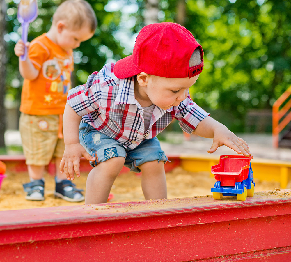 A young boy is playing in a sandbox with a toy truck, deeply immersed in his play while another child is in the background.