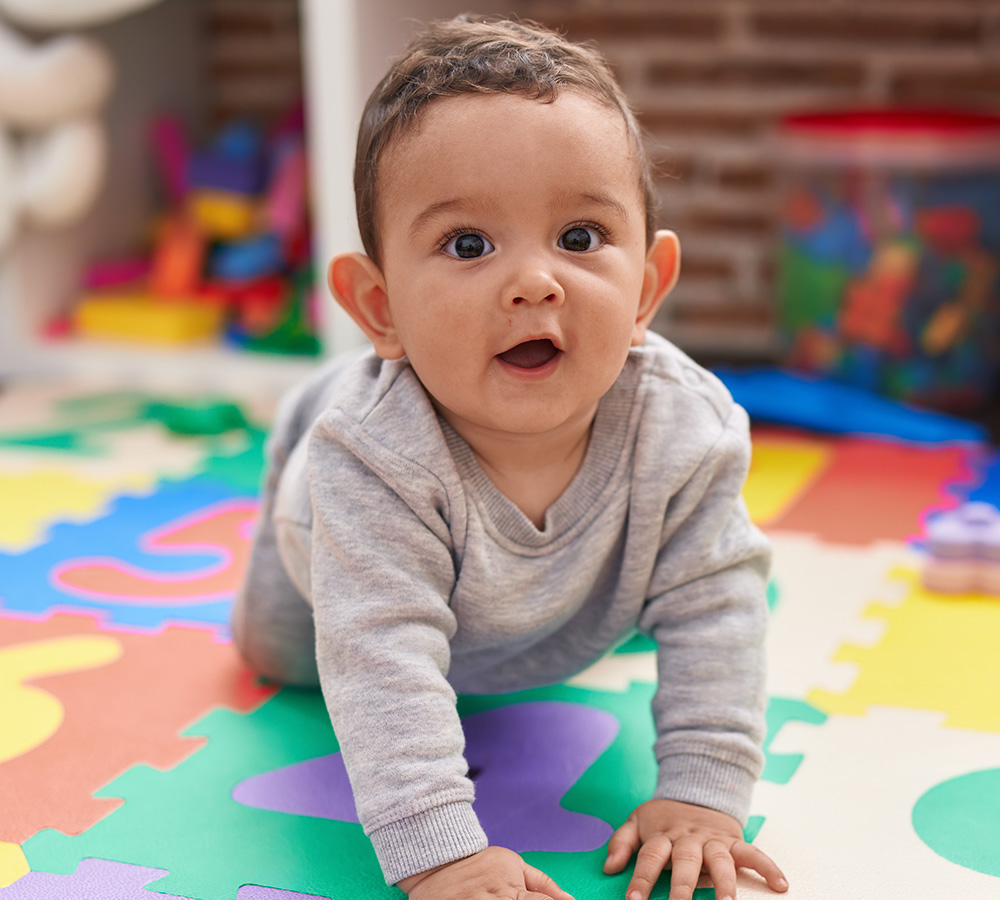 A baby is crawling on a colorful mat, looking surprised and happy, with a playful background filled with toys.
