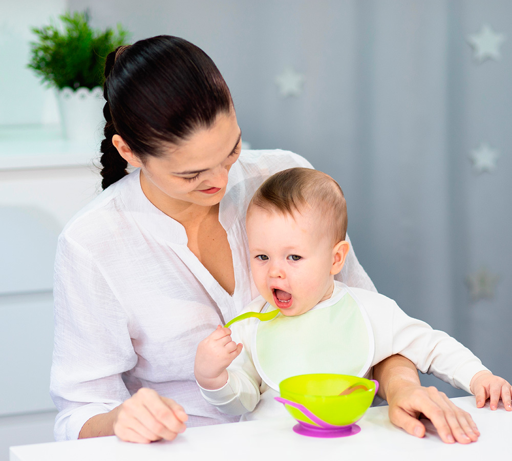 A teacher is feeding a baby, who is making a playful face while holding a spoon, with a bright and cozy background.