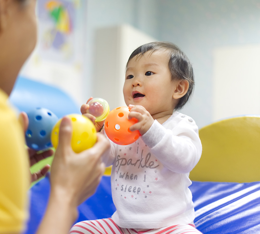 A baby joyfully holding colorful balls, looking excited and happy in a playful environment, with an teacher nearby.