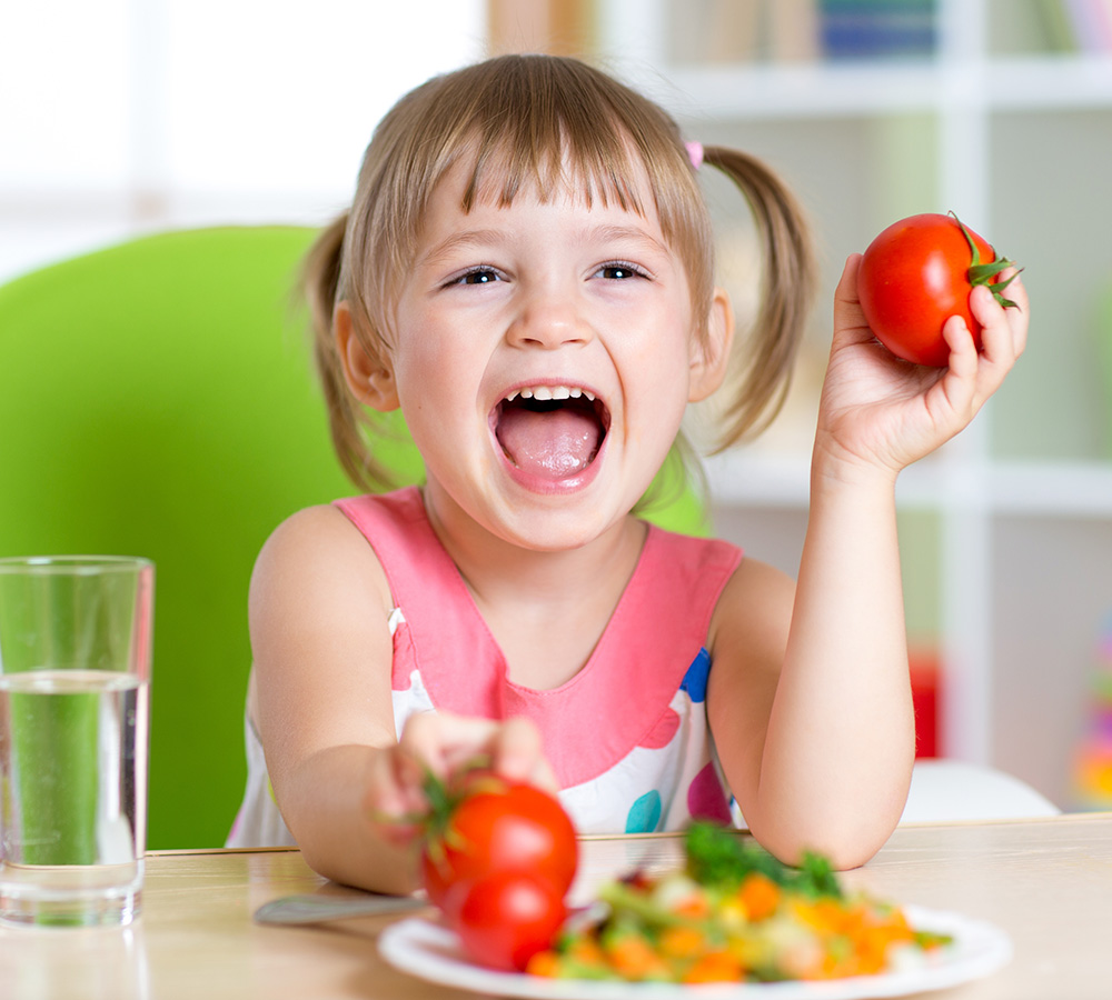 A joyful girl smiles widely while holding a tomato, surrounded by a colorful plate of food.