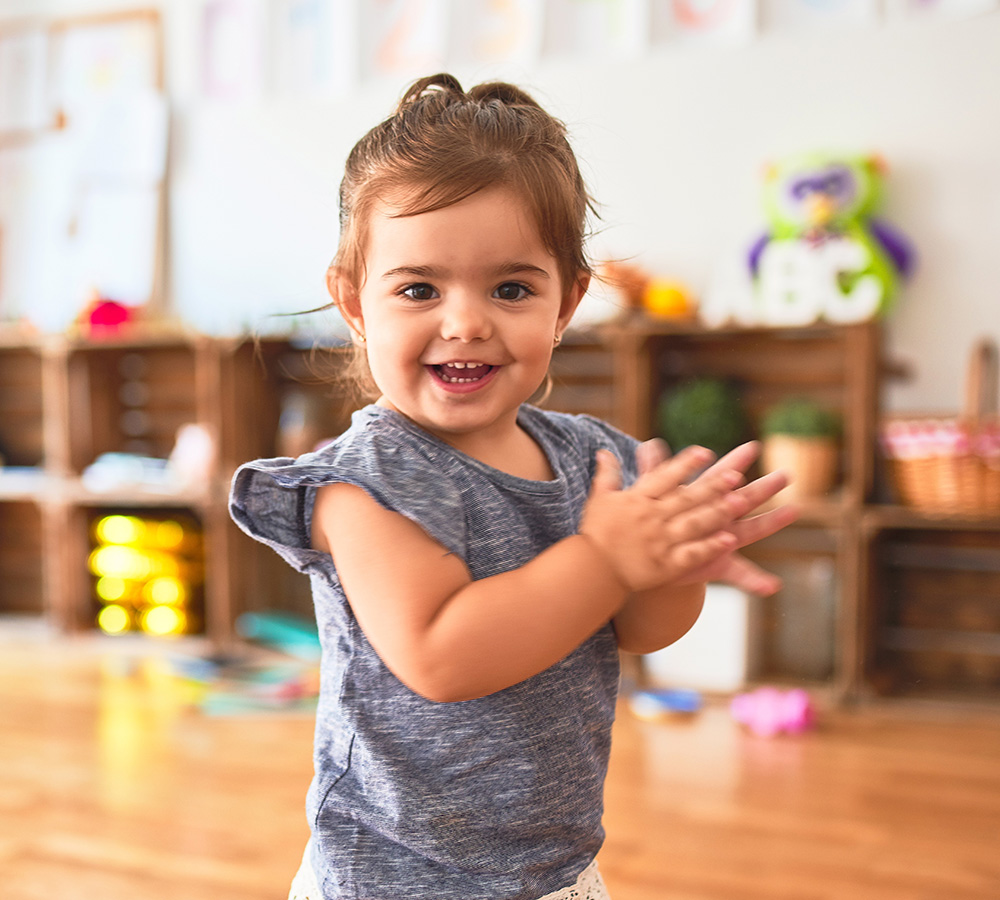 A young girl is joyfully clapping her hands, radiating happiness in a playful environment filled with toys.