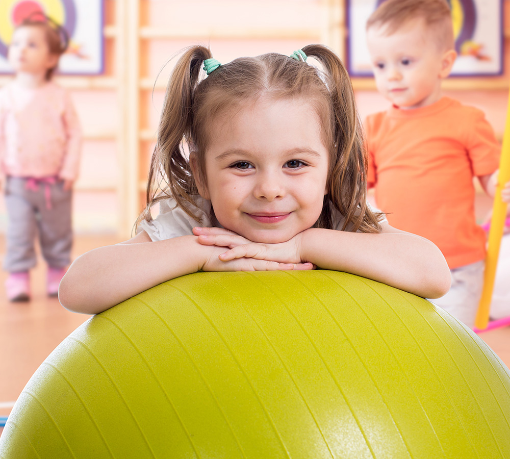 A happy girl rests on a yellow ball, smiling, with playful children in the background.